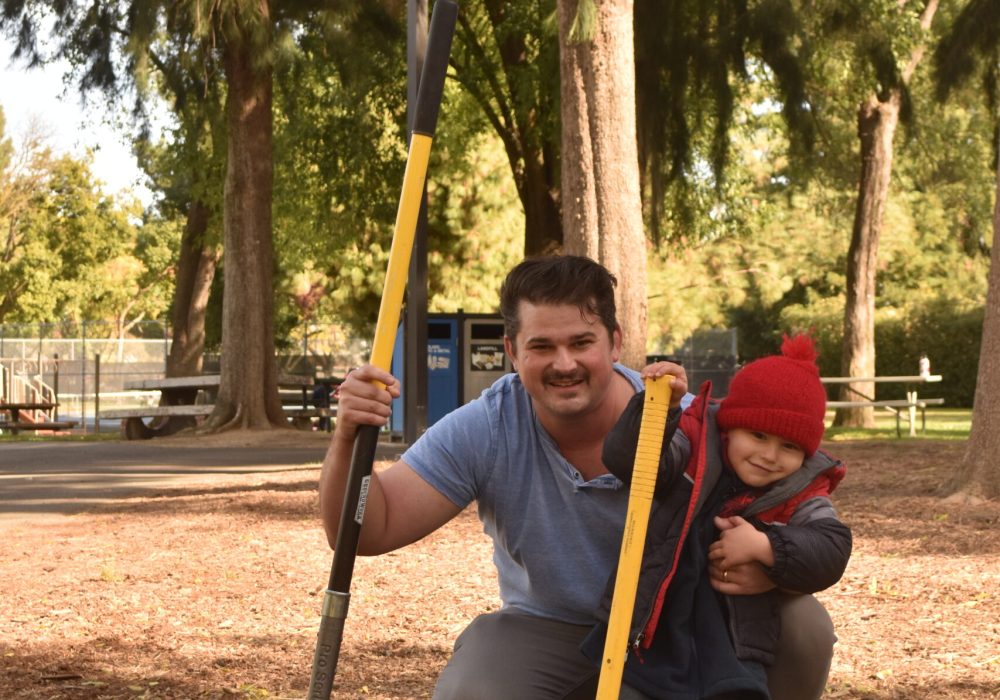 Man and child pose with planting tools at a park.