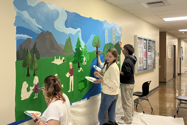 Students paint a mural of grass, mountains, trees, people, river, and bridge.